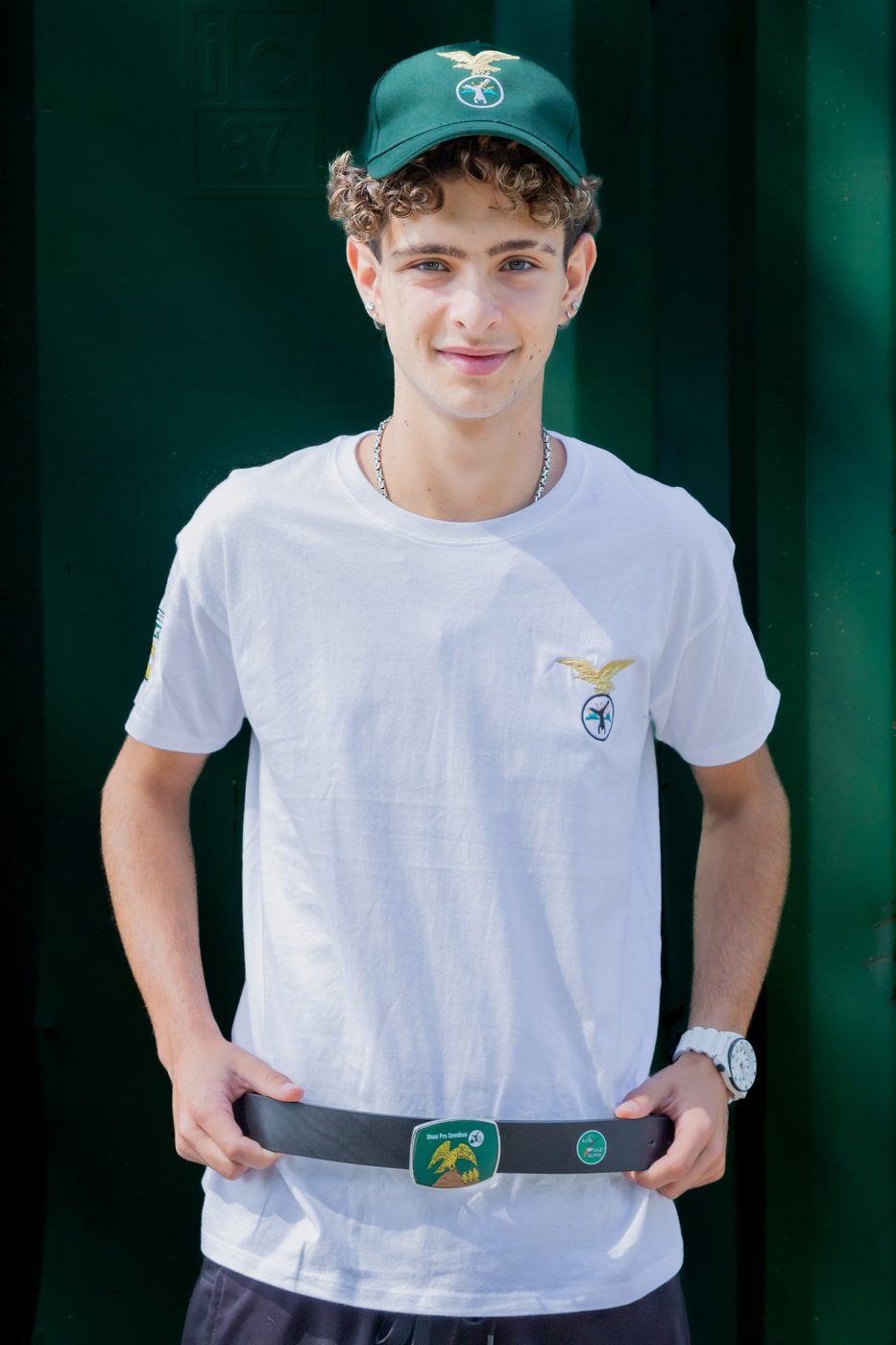 Ragazzo con cintura con fibbia decorata ad aquila, maglietta bianca e cappellino verde